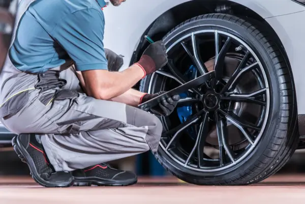 A mechanic in gray overalls and work gloves kneels beside a white car's front wheel, holding a pen and clipboard while inspecting the tire and blue brake caliper.