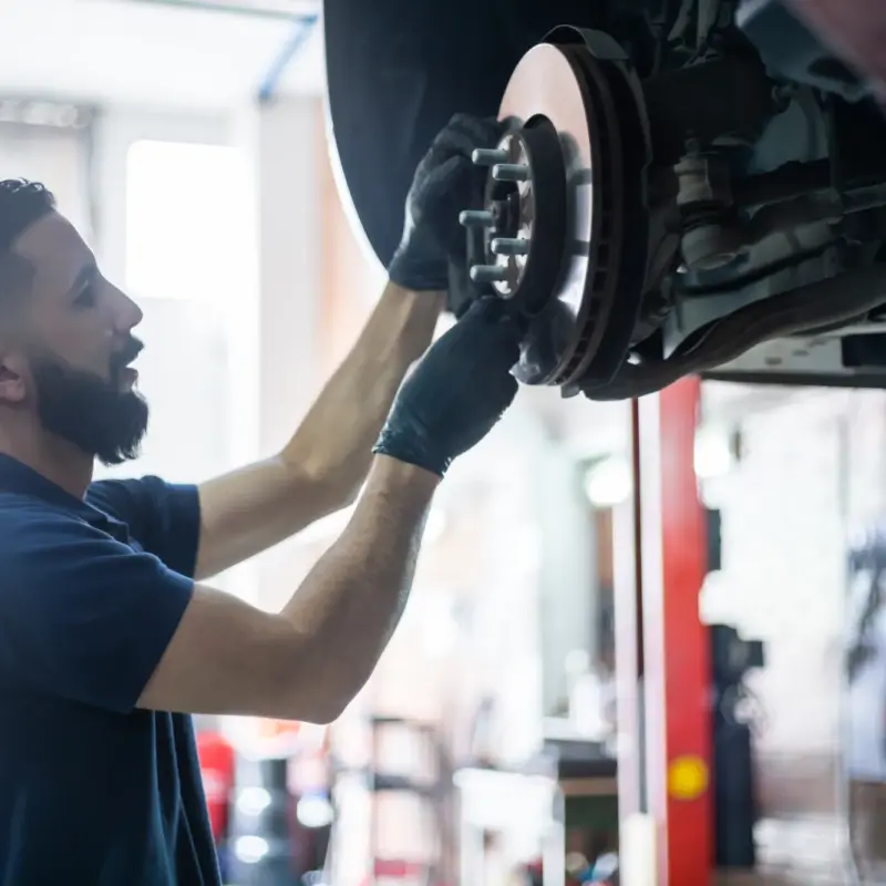A mechanic wearing black gloves inspects the brake rotor and wheel hub of a car lifted in a repair shop.