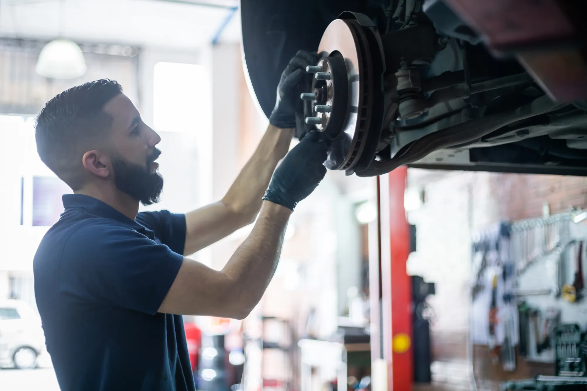 A mechanic wearing black gloves inspects the brake rotor and wheel hub of a car lifted in a repair shop.