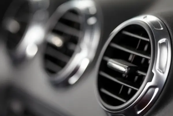 Close-up of a round chrome-trimmed air conditioning vent on a black car dashboard, with a second vent blurred in the background.