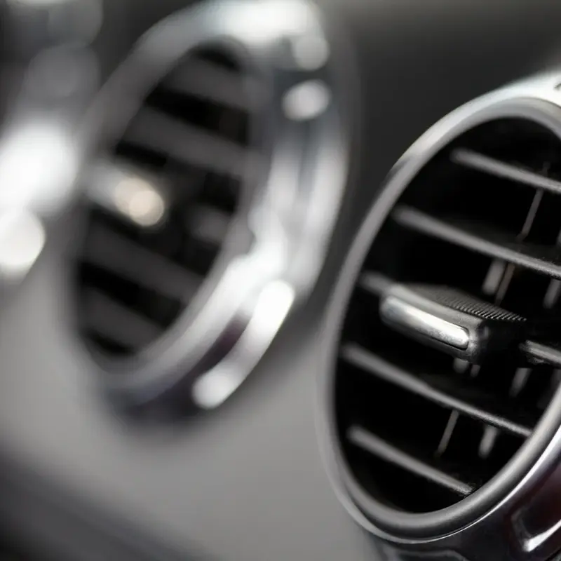 Close-up of a round chrome-trimmed air conditioning vent on a black car dashboard, with a second vent blurred in the background.