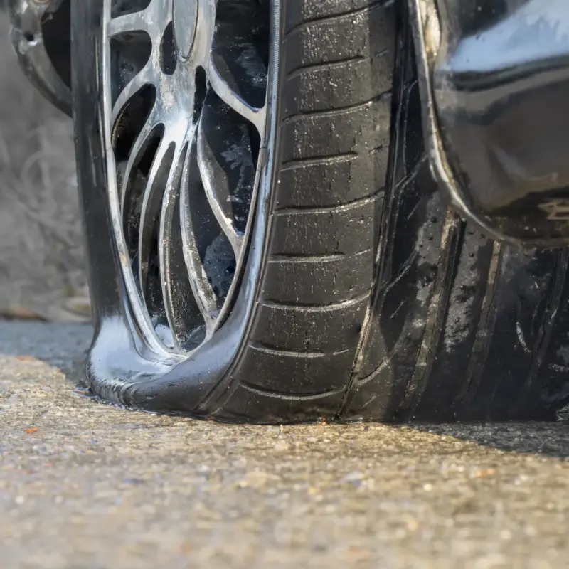 Close-up of a flat, deflated tire on an alloy wheel of a dark car resting on wet asphalt.